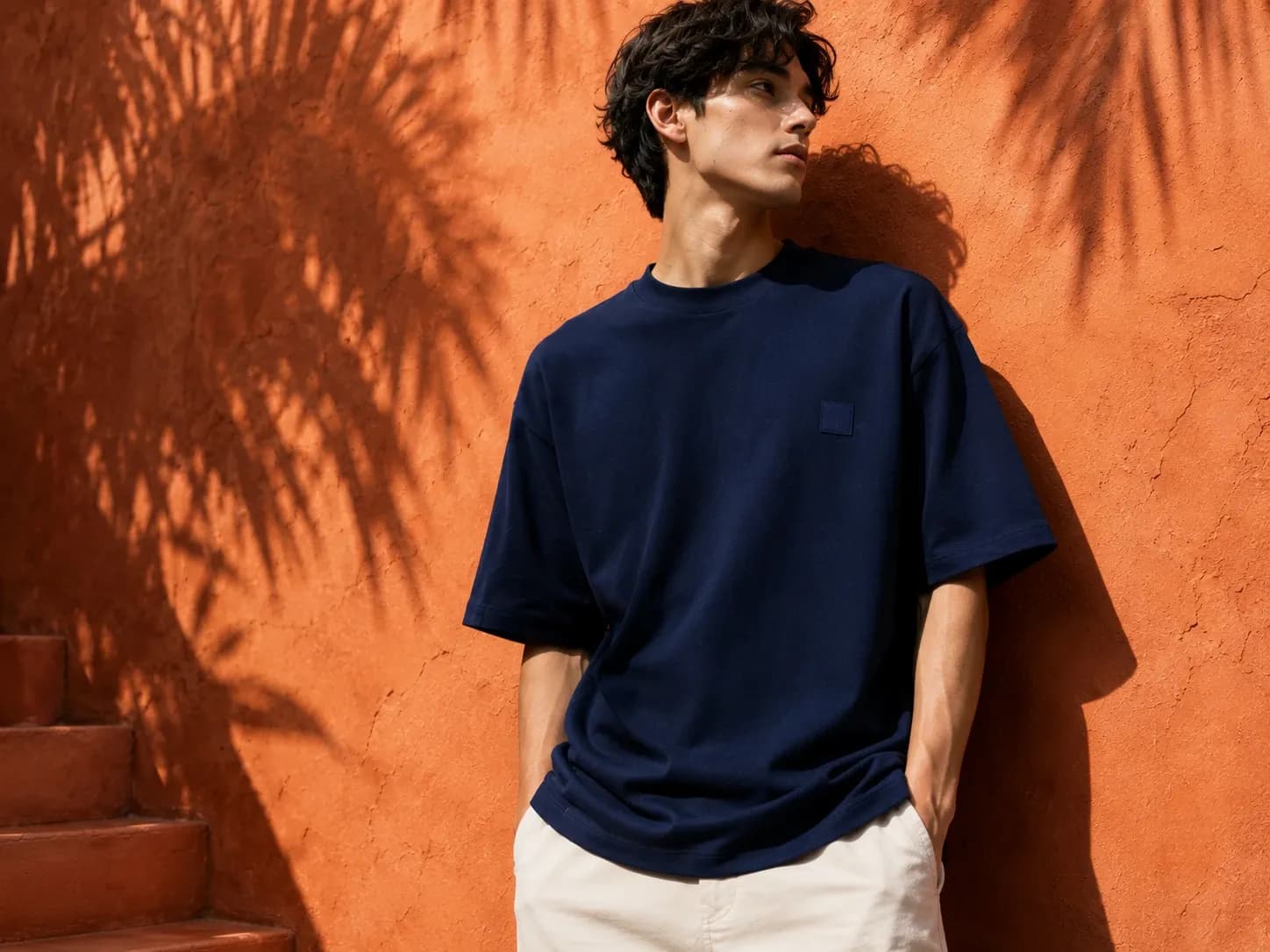 A young man in a navy oversized t-shirt and cream trousers leans against a terracotta wall with palm shadows, looking to the side
